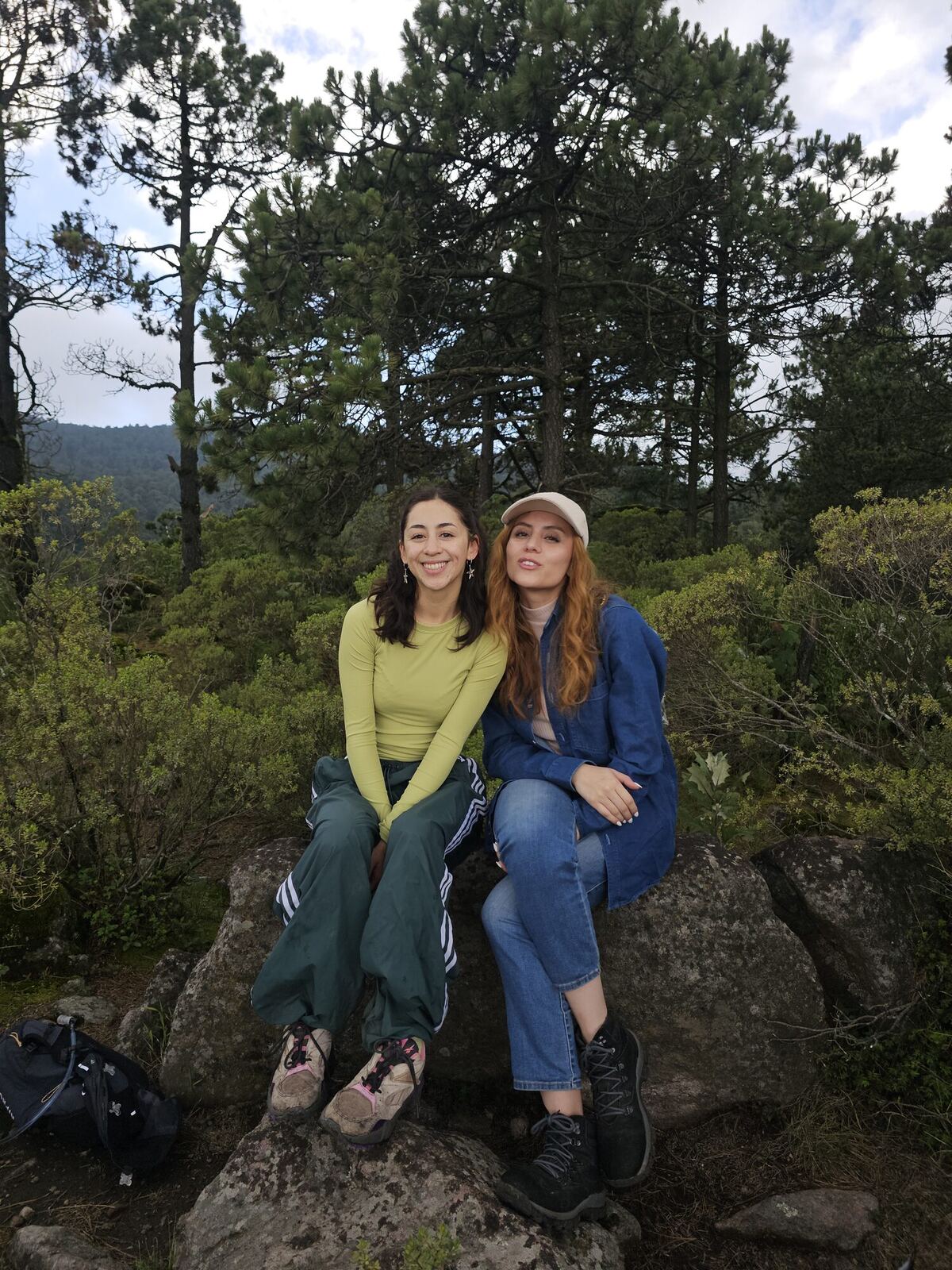 Dos amigas sonriendo sentadas en una roca entre pinos