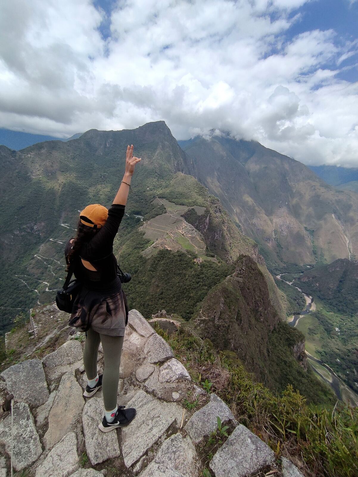 Una mujer con el brazo en alto en Huayna Picchu, con Machu Picchu abajo